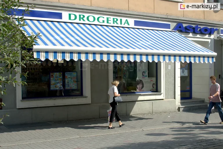 widok na witrynę sklepową placówki handlowej o nazwie „DROGERIA”, nad którą zamontowano dużą markizę tarasową w klasyczne, pionowe pasy. Poszycie markizy składa się z naprzemiennych pasów w kolorze białym oraz intensywnie niebieskim. Krawędź markizy wykończona jest ozdobną, falowaną falbaną, na której powtórzono ten sam wzór kolorystyczny. Konstrukcja wsporcza i czołowa listwa markizy wykonane są w kolorze białym. Nad markizą widnieje szyld z zielonym napisem „DROGERIA” na białym tle, ujęty w niebieskie obramowanie. Obok znajduje się fragment kolejnej witryny z niebieskim napisem „Astor”. W oknach wystawowych drogerii widoczne są plakaty reklamowe produktów kosmetycznych oraz asortyment sklepowy. Przed budynkiem przebiega chodnik wyłożony szarą kostką brukową, po którym poruszają się przechodnie: kobieta w białej bluzce i ciemnych spodniach niosąca torbę oraz mężczyzna w pasiastej koszulce i jeansach. Elewacja budynku jest w stonowanym, beżowo-szarym kolorze. Markiza pełni funkcję zarówno praktyczną, chroniąc wnętrze sklepu przed nadmiernym nagrzewaniem i promieniowaniem UV, jak i estetyczną, wyróżniając lokal na tle pierzei ulicznej.