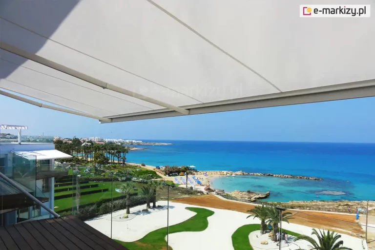 Zdjęcie przedstawia widok z wysokiego tarasu lub balkonu na malownicze wybrzeże morskie, przesłonięty od góry rozłożystą markizą tarasową. Markiza posiada poszycie w kolorze białym, które jest rozpięte na maksymalny możliwy wysięg. Konstrukcja w kolorze białym jest zamontowana do elewacji nowoczesnego budynku z przeszklonymi balustradami. Poniżej rozpościera się widok na luksusowy kurort z intensywnie zielonymi, tarasowymi trawnikami, palmami oraz jasnymi alejkami o finezyjnych kształtach. W oddali widoczna jest piaszczysta plaża z niebieskimi parasolami oraz skalista zatoka z krystalicznie czystą wodą o turkusowym odcieniu. Linia horyzontu oddziela spokojną taflę morza od bezchmurnego, błękitnego nieba. Na dalszym planie wzdłuż linii brzegowej ciągnie się zabudowa miejska z niskimi, jasnymi budynkami. Perspektywa zdjęcia z poziomu tarasu podkreśla funkcjonalność markizy jako elementu zapewniającego komfortowy cień w nasłonecznionym, śródziemnomorskim klimacie, nie ograniczając przy tym panoramicznego widoku na krajobraz.