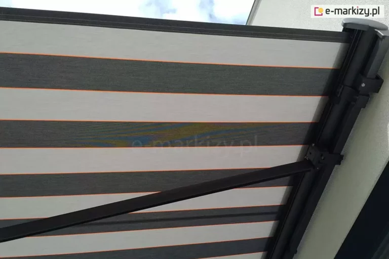 Markiza Palladio o dużym wysięgu z maksymalnie rozłożonymi ramionami. Po prawej widoczne mocowanie aluminiowej kasety w kolorze srebrzysty grafit do białej ściany. Nad ramieniem markizy widoczne tkaninowe poszycie w pady o dwóch odcieniach szarości.