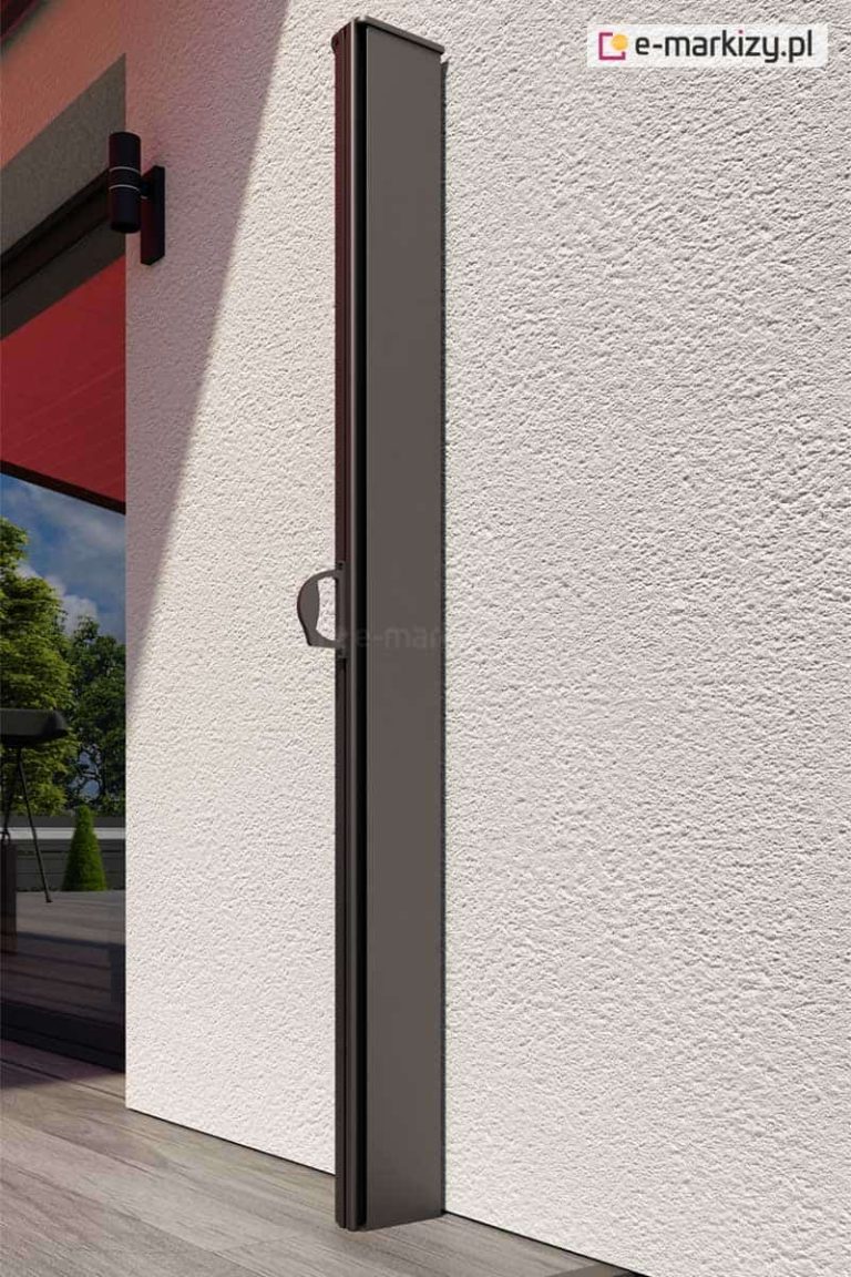 Zamocowana do ściany o jasnej elewacji z tynkiem strukturalnym pionowa kaseta markizy bocznej Bora w kolorze antracytowym. Tkanina pozostaje szczelnie zamknięta w kasecie. W głębi widoczna wnęka okienna, w której odbija się zieleń ogrodu. W pobliżu narożnika wnęki zamocowana nowoczesna lampa fasadowa w czarnym kolorze.