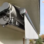 Podwieszona za pomocą uchwytów sufitowych do płyty balkonu wyższego piętra markiza Australia w kolorze srebrzysty grafit z widocznym zaczepem awaryjnego otwierania oraz przewodem zasilającym silnika. Zwinięta belka przednia pozwala dostrzec prostą falbanę wykonaną z jasnoszarej tkaniny. W tle za balkonem, do którego przymocowano markizę, widoczne drzewa oraz niebieskie niebo.