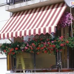 Klasyczny balkon w bloku mieszkalnym, udekorowany obfitymi kompozycjami kwiatowymi i osłonięty tradycyjną markizą balkonową z ramionami poręczowymi. Markiza posiada poszycie w szerokie, pionowe pasy w kolorach bordowym i kremowym, wykończone na dole dekoracyjną, pofalowaną falbaną z białą lamówką. Materiał jest rozwinięty pod skosem, tworząc stabilne zadaszenie nad całą szerokością balkonu. Balustrada balkonu jest niemal całkowicie zasłonięta przez gęste, zwisające pelargonie o intensywnie czerwonych i różowych kwiatach, które rosną w podłużnych skrzynkach. Dodatkowo, w prawym górnym rogu pod sufitem wnęki, zawieszona jest donica z bujną, różową surfinią. Balustrada posiada wypełnienie z półtransparentnych, żółtawych paneli, a po prawej stronie widoczny jest pionowy fragment rynny deszczowej oraz zamontowana na elewacji antena satelitarna. Ściana budynku jest biała, o gładkiej strukturze, co stanowi jasne tło dla nasyconych barw kwiatów i pasiastej tkaniny osłonowej. Całość przedstawia przytulną i zadbaną przestrzeń rekreacyjną w pełnym świetle dziennym.