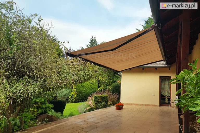 Zdjęcie przedstawia rozłożoną poziomą markizę tarasową Presto zamontowaną do konstrukcji dachu jednorodzinnego budynku. Poszycie markizy wykonane jest z materiału w ciepłym, beżowym lub piaskowym kolorze z delikatnymi, ciemniejszymi pasami biegnącymi równolegle do krawędzi bocznych. Tkanina jest mocno napięta na ciemnobrązowym, aluminiowym stelażu z widocznymi ramionami przegubowymi, które podtrzymują konstrukcję od spodu. Markiza jest w pełni wysunięta, osłaniając znaczną część przestronnego tarasu wyłożonego jasnobrązowymi, matowymi płytkami ceramicznymi. Taras przylega do ściany budynku o gładkiej elewacji w kolorze kremowym lub bardzo jasnym żółtym. Po prawej stronie widoczne są przeszklone drzwi tarasowe z brązową ramą, przez które dostrzec można jasną firankę wewnątrz pomieszczenia. Obok drzwi stoi białe, składane krzesło ogrodowe. Wzdłuż krawędzi tarasu, na granicy z ogrodem, ustawiona jest niska donica z pomarańczowymi kwiatami. Tło zdjęcia stanowi gęsta, zadbana roślinność ogrodowa z przewagą wysokich krzewów liściastych po lewej stronie oraz formowanych iglaków w głębi. Pomiędzy krzewami widoczny jest fragment przystrzyżonego, zielonego trawnika. Niebo nad ogrodem jest jasne, lekko zachmurzone, co sugeruje słoneczny, ale nie upalny dzień. Całość kompozycji ukazuje praktyczne zastosowanie markizy Presto jako elementu chroniącego strefę wypoczynkową przed słońcem w otoczeniu przydomowej zieleni.
