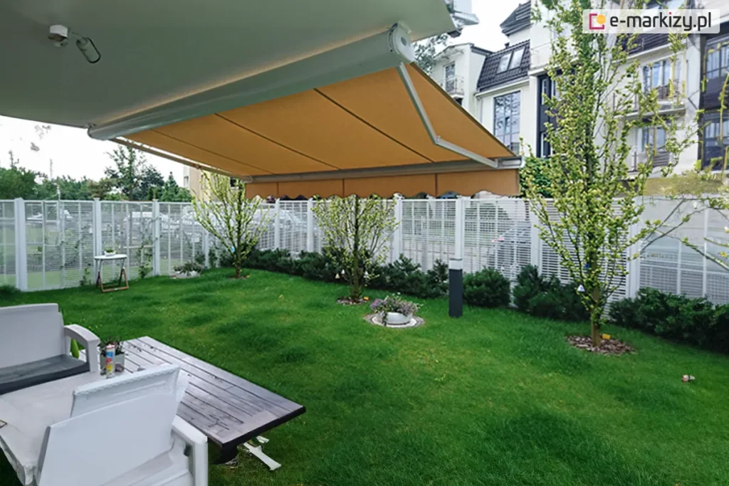 Zdjęcie wykonane z perspektywy ogrodu przedstawia w pełni rozłożoną markizę tarasową zamontowaną do elewacji nowoczesnego budynku. Poszycie markizy wykonane jest z gładkiego materiału w ciepłym, musztardowym lub miodowym kolorze, zakończone klasyczną, falowaną falbaną. Konstrukcja wsparta jest na przegubowych, białych ramionach wykonanych z aluminium, które harmonizują z jasnym kolorem rury nawojowej. Pod zadaszeniem znajduje się strefa wypoczynkowa z drewnianym stołem o ciemnym blacie oraz białymi fotelami z szarymi poduszkami. Tło stanowi zadbany, zielony trawnik z młodymi drzewkami liściastymi oraz niskimi krzewami iglastymi nasadzonymi wzdłuż białego, panelowego ogrodzenia. W oddali widać wielorodzinną zabudowę miejską o białych elewacjach z licznymi przeszkleniami oraz fragmenty błękitnego nieba.
