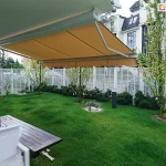 Zdjęcie wykonane z perspektywy ogrodu przedstawia w pełni rozłożoną markizę tarasową zamontowaną do elewacji nowoczesnego budynku. Poszycie markizy wykonane jest z gładkiego materiału w ciepłym, musztardowym lub miodowym kolorze, zakończone klasyczną, falowaną falbaną. Konstrukcja wsparta jest na przegubowych, białych ramionach wykonanych z aluminium, które harmonizują z jasnym kolorem rury nawojowej. Pod zadaszeniem znajduje się strefa wypoczynkowa z drewnianym stołem o ciemnym blacie oraz białymi fotelami z szarymi poduszkami. Tło stanowi zadbany, zielony trawnik z młodymi drzewkami liściastymi oraz niskimi krzewami iglastymi nasadzonymi wzdłuż białego, panelowego ogrodzenia. W oddali widać wielorodzinną zabudowę miejską o białych elewacjach z licznymi przeszkleniami oraz fragmenty błękitnego nieba.