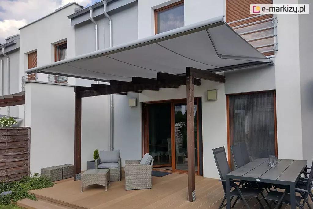 Niewielka drewniana pergola ustawiona na tarasie z miejscami do siedzenia z niskim stolikiem oraz dużym stołem jadalnianym w kolorze czarnym z czarnymi krzesłami osłonięta została od góry zamocowaną do ściany o jasnej elewacji markizą Presto PLUS ze srebrną konstrukcją i jasnoszarym poszyciem. W tle pod markizą widoczne otwarte okno z wyjściem na taras oraz okno osłonięte w całości moskitierą.
