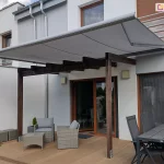 Niewielka drewniana pergola ustawiona na tarasie z miejscami do siedzenia z niskim stolikiem oraz dużym stołem jadalnianym w kolorze czarnym z czarnymi krzesłami osłonięta została od góry zamocowaną do ściany o jasnej elewacji markizą Presto PLUS ze srebrną konstrukcją i jasnoszarym poszyciem. W tle pod markizą widoczne otwarte okno z wyjściem na taras oraz okno osłonięte w całości moskitierą.