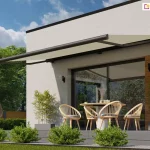 Zdjęcie prezentuje nowoczesną markizę przeciwsłoneczną Casablanca Aluprof w pełnym rozłożeniu, zamontowaną na białej elewacji budynku o minimalistycznej architekturze. Markiza przeciwsłoneczna Casablanca posiada poszycie w kolorze piaskowym lub beżowym, które rzuca głęboki, kojący cień na strefę wypoczynkową tarasu. Konstrukcja kasetowa oraz ramiona przegubowe urządzenia wykonane są z aluminium lakierowanego na matowy kolor antracytowy, co tworzy elegancki kontrast z jasną ścianą i drewnianą szalówką dekoracyjną. Pod markizą znajduje się zaaranżowana przestrzeń jadalniana z okrągłym stołem oraz czterema ażurowymi krzesłami z jasnego drewna, a obok widoczny jest fragment plecionego fotela wiszącego typu kokon. Na pierwszym planie, wzdłuż krawędzi tarasu wykonanego z szarych desek kompozytowych, posadzone są w żwirze niskie, zielone krzewy ozdobne o kulistym pokroju. W tle rozciąga się soczysty trawnik oraz gęsta ściana wysokich, zielonych drzew liściastych pod jasnym, lekko zachmurzonym niebem. Po prawej stronie kadru widoczny jest również fragment drewnianego stelaża hamaka, co podkreśla rekreacyjny charakter miejsca. Przeszklone drzwi tarasowe odbijają otaczający ogród, a czarne, pionowe kinkiety na elewacji dopełniają nowoczesny design bryły domu.