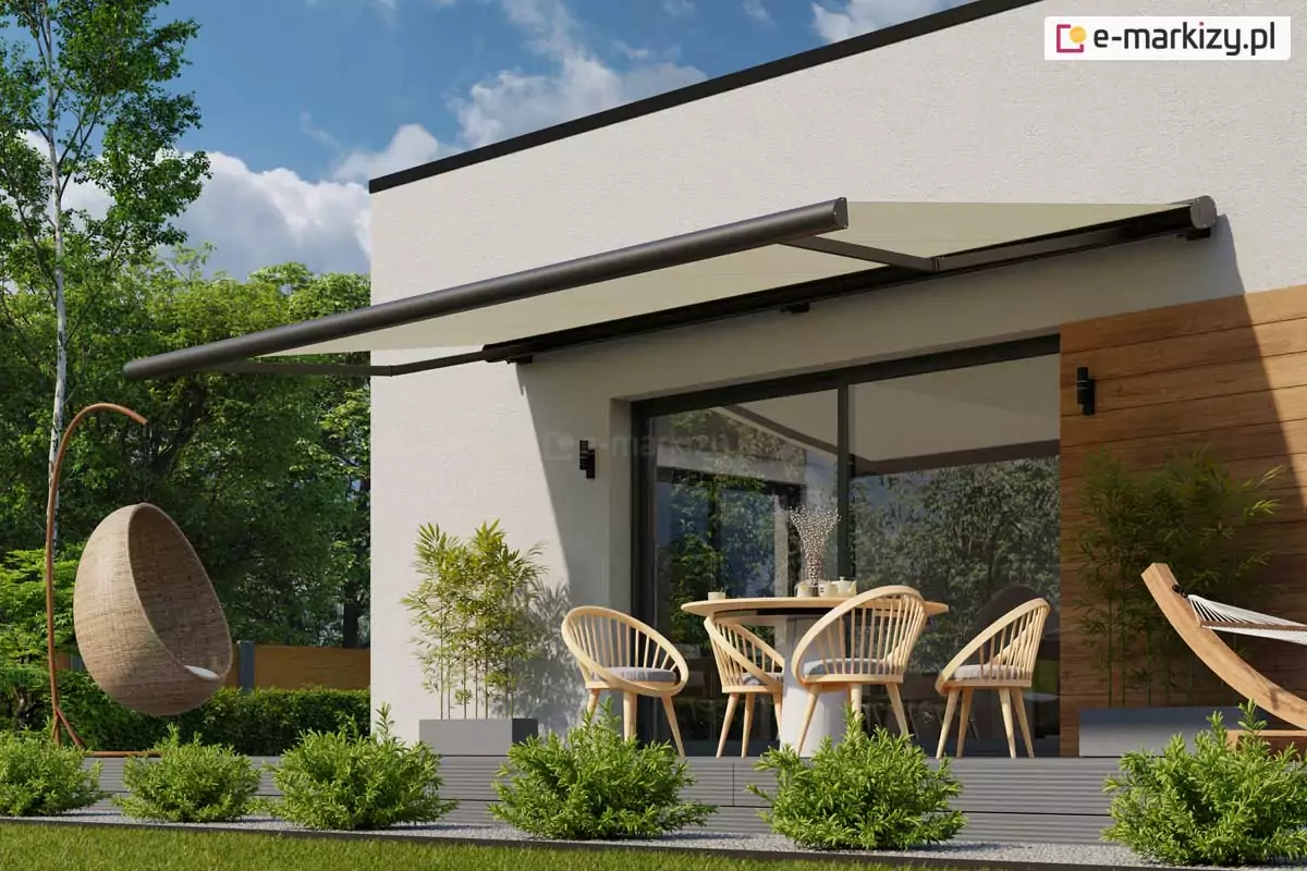 Zdjęcie prezentuje nowoczesną markizę przeciwsłoneczną Casablanca Aluprof w pełnym rozłożeniu, zamontowaną na białej elewacji budynku o minimalistycznej architekturze. Markiza przeciwsłoneczna Casablanca posiada poszycie w kolorze piaskowym lub beżowym, które rzuca głęboki, kojący cień na strefę wypoczynkową tarasu. Konstrukcja kasetowa oraz ramiona przegubowe urządzenia wykonane są z aluminium lakierowanego na matowy kolor antracytowy, co tworzy elegancki kontrast z jasną ścianą i drewnianą szalówką dekoracyjną. Pod markizą znajduje się zaaranżowana przestrzeń jadalniana z okrągłym stołem oraz czterema ażurowymi krzesłami z jasnego drewna, a obok widoczny jest fragment plecionego fotela wiszącego typu kokon. Na pierwszym planie, wzdłuż krawędzi tarasu wykonanego z szarych desek kompozytowych, posadzone są w żwirze niskie, zielone krzewy ozdobne o kulistym pokroju. W tle rozciąga się soczysty trawnik oraz gęsta ściana wysokich, zielonych drzew liściastych pod jasnym, lekko zachmurzonym niebem. Po prawej stronie kadru widoczny jest również fragment drewnianego stelaża hamaka, co podkreśla rekreacyjny charakter miejsca. Przeszklone drzwi tarasowe odbijają otaczający ogród, a czarne, pionowe kinkiety na elewacji dopełniają nowoczesny design bryły domu.