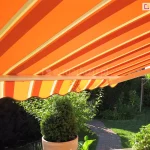 Zdjęcie prezentuje zbliżenie na rozłożoną markizę Mezzo mol, ukazaną z perspektywy osoby znajdującej się bezpośrednio pod jej poszyciem. Materiał markizy charakteryzuje się wzorem w szerokie, pionowe pasy w intensywnych kolorach pomarańczowym i czerwonym, które są przedzielone cieńszymi, jasnobeżowymi liniami. Światło słoneczne przenikające przez tkaninę podkreśla nasycenie barw i tworzy ciepłą aurę w zacienionej strefie. Konstrukcja wspierająca markizę Mezzo mol posiada biały profil przedni, do którego zamocowana jest ozdobna, falowana falbana, powtarzająca kolorystykę i wzór głównej części poszycia. W tle, pod osłoną markizy, rozpościera się widok na zadbany ogród z zielonym trawnikiem i krętą ścieżką ułożoną z kostki brukowej w odcieniach czerwieni i szarości. Na pierwszym planie widoczne są dwa formowane w kształt stożka krzewy iglaste, umieszczone w dużych, jasnych, ceramicznych donicach. W dalszej części ogrodu znajdują się gęste krzewy liściaste, wysoki żywopłot oraz drewniana konstrukcja huśtawki ogrodowej. Całość obrazu ilustruje efektywność markizy Mezzo mol w tworzeniu komfortowej, zacienionej przestrzeni do wypoczynku na świeżym powietrzu, chroniącej przed bezpośrednim promieniowaniem słonecznym.