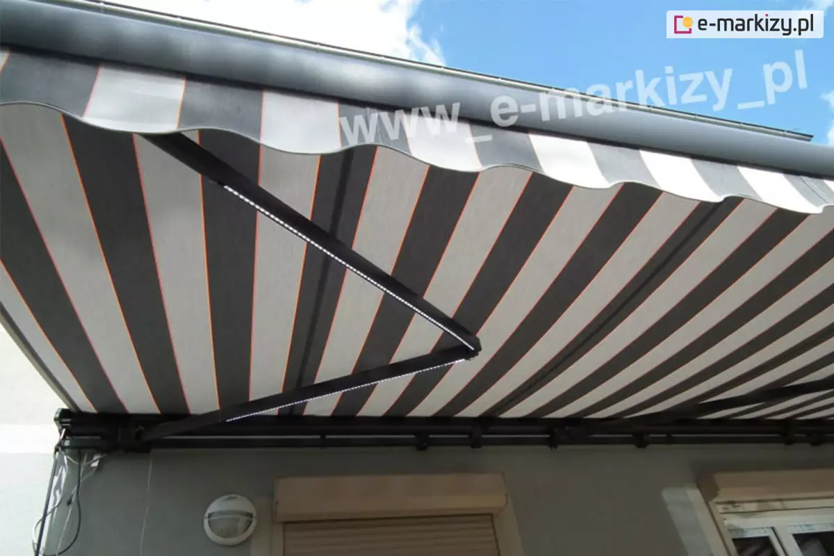 Zdjęcie prezentuje frontowe ujęcie od dołu markizy tarasowej rozłożonej nad elewacją nowoczesnego budynku. Markiza przeciwsłoneczna Palladio charakteryzuje się poszyciem w szerokie, pionowe pasy w odcieniach grafitu i jasnej szarości, które są przedzielone cienkimi prążkami w kolorze neonowej pomarańczy. Konstrukcja markizy, w tym profil czołowy oraz ramiona przegubowe, została wykonana z aluminium lakierowanego na kolor ciemnoantracytowy. Na ramionach przegubowych zamontowano oświetlenie LED w formie liniowych listew, które emitują białe, zimne światło, doświetlając przestrzeń tarasową. Markiza posiada ozdobną, falowaną falbanę z lamówką, która estetycznie wykańcza profil czołowy. W tle widoczna jest jasnoszara ściana budynku z zamontowaną okrągłą lampą elewacyjną oraz fragmentami rolet zewnętrznych nad oknami. Górną część kadru wypełnia błękitne niebo z białymi, puszystymi chmurami, co podkreśla letni i funkcjonalny charakter prezentowanej osłony przeciwsłonecznej.