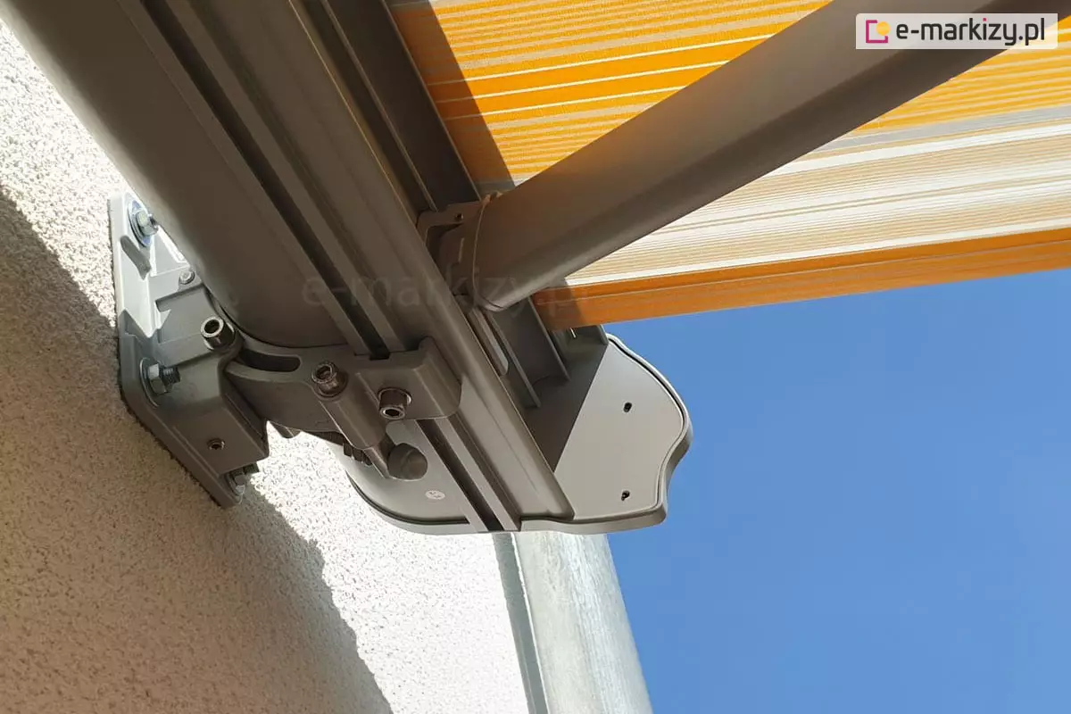 Zamocowany do ściany uchwyt konsolowy markizy Dakar ze zbliżeniem miejsca jego łączenia z kasetą markizy. W tle widoczne mocowanie ramienia w kasecie, zaślepka boczna, fragment poszycia w żółte i białe pasy, biała ściana i niebieskie niebo.