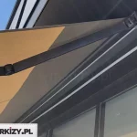 Zdjęcie prezentuje nowoczesną markizę ścienną zainstalowaną na ciemnej elewacji budynku, przedstawioną z perspektywy od dołu, co eksponuje jej mechanizm i ramię przegubowe. Markiza Moderno posiada konstrukcję w kolorze antracytowym o matowym wykończeniu, a jej ramię jest w pełni wyprostowane, podtrzymując napięte, beżowe poszycie o wyraźnym splocie tkaniny. Materiał markizy rzuca geometryczny cień na ciemną ścianę oraz dużą szklaną witrynę znajdującą się poniżej zadaszenia. System mocowania ramienia do profilu nośnego charakteryzuje się minimalistyczną, kanciastą formą, typową dla współczesnej architektury. W lewym górnym rogu kadru widoczny jest fragment czystego, błękitnego nieba, co podkreśla funkcję ochronną produktu w słoneczny dzień.