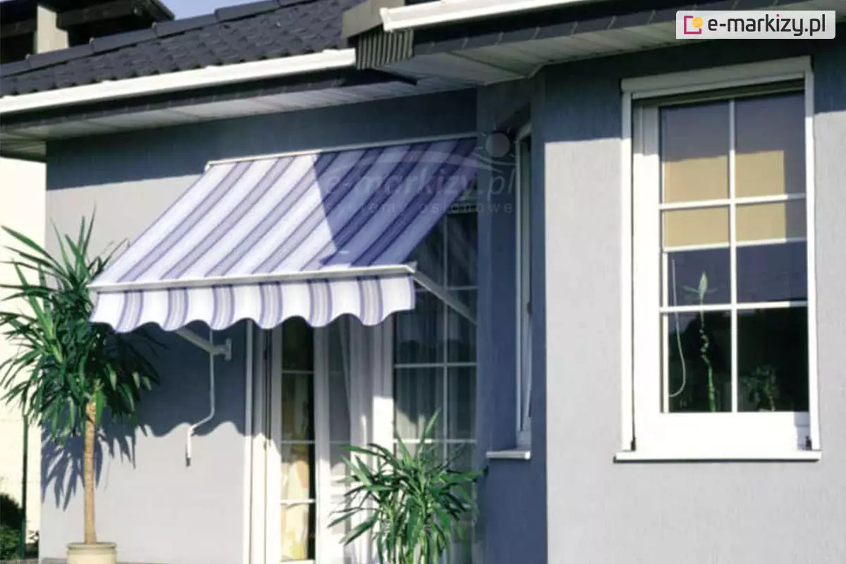 Zdjęcie prezentuje instalację zewnętrznej markizy balkonowo-sklepowej Italia w kasecie na elewacji domu jednorodzinnego. Budynek posiada ściany wykończone tynkiem w kolorze gołębim lub jasnoszarym oraz dach pokryty ciemną dachówką z białą podbitką i rynnami. Markiza jest zamontowana nad przeszklonymi drzwiami tarasowymi. Jej poszycie charakteryzuje się wzorem w pionowe pasy w odcieniach bieli, błękitu i fioletu, a krawędź wykończona jest ozdobną, falowaną falbaną. Konstrukcja osłony opiera się na białych ramionach bocznych, które są przytwierdzone do ściany budynku poniżej poziomu kasety. Przy lewym ramieniu zwisa także korba do ręcznego sterowania markizą. Obok wejścia na taras, po lewej stronie, ustawiona jest wysoka roślina doniczkowa o długich, zielonych liściach przypominająca jukę lub dracenę. Po prawej stronie widoczne jest mniejsze okno z białymi szprosami, w którym zamontowana jest wewnętrzna roleta materiałowa w kolorze beżowym. Całość kompozycji utrzymana jest w jasnej kolorystyce, w pełnym świetle dziennym, co podkreśla funkcjonalność markizy jako ochrony przeciwsłonecznej dla strefy wejściowej.