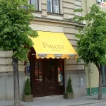 Markiza Italia bez kasety z ramionami do ściany o żółtym poszyciu z dużym nadrukiem logo nad belką przednią i falowanej falbanie z nadrukami logo producenta piwa zamontowana została na betonowej fasadzie budynku nad drewnianymi drzwiami wejścia do kawiarni usytuowanej w narożniku budynku, przed którym posadzono dwa drzewa liściaste i ustawiono kilka donic z roślinkami.