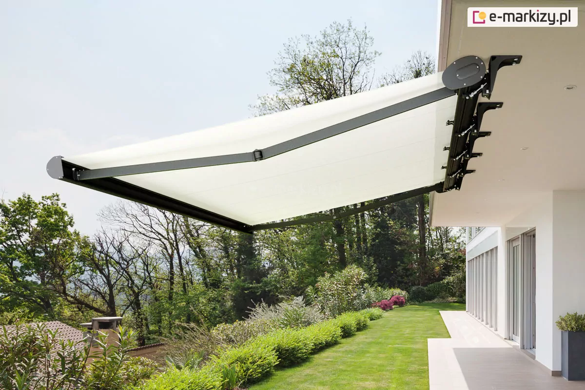 Zdjęcie prezentuje nowoczesną markizę Casablanca zamontowaną pod podbitką dachu białego budynku o minimalistycznej architekturze. Markiza Casablanca charakteryzuje się pełną kasetą o opływowym kształcie, która w tym wariancie posiada konstrukcję w kolorze ciemnego antracytu lub czerni, co wyraźnie odcina się na tle jasnej elewacji. Poszycie markizy wykonane jest z gładkiego, jasnego materiału w odcieniu bieli lub bardzo jasnego kremu, który po całkowitym rozwinięciu tworzy dużą płaszczyznę ochrony przed słońcem. Konstrukcja wspierająca materiał opiera się na dwóch przegubowych ramionach o kanciastym profilu, które są wysunięte do przodu, zapewniając stabilność i odpowiednie napięcie tkaniny. System mocowania markizy Casablanca obejmuje szereg solidnych uchwytów przytwierdzonych bezpośrednio do dolnej części zadaszenia budynku. W tle widoczny jest rozległy ogród z soczystą, zieloną trawą oraz gęstym pasem niskich krzewów i roślinności ozdobnej, za którymi rozciąga się ściana wysokich liściastych drzew. Po prawej stronie zdjęcia znajduje się taras wyłożony jasnymi płytkami, który biegnie wzdłuż przeszklonej ściany parteru domu, a na jego krawędzi stoi nowoczesna, ciemna donica z rośliną. Perspektywa zdjęcia ujęta jest od dołu, co podkreśla techniczne aspekty budowy markizy oraz jej duży wysięg nad strefą wypoczynkową. Niebo jest jasne i lekko zamglone, co sugeruje słoneczny, ale nie upalny dzień, idealny do korzystania z osłony tarasowej.