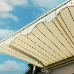 Zbliżenie na krawędź przednią i spód poszycia markizy Adagio, ukazujące detale wykończenia tkaniny oraz konstrukcję falbany. Materiał markizy ma kolor kremowy lub jasnobeżowy z subtelnymi, ciemniejszymi pionowymi pasami o różnej szerokości. Na krawędzi czołowej znajduje się dekoracyjna falbana o klasycznym, falistym kształcie, która została obszyta ciemniejszą lamówką dla wzmocnienia brzegu i podkreślenia formy. Tkanina jest precyzyjnie napięta na aluminiowym profilu przednim w kolorze białym. Pod poszyciem widoczny jest fragment białego ramienia przegubowego oraz rury nawojowej markizy Adagio, zamontowanej na jasnej elewacji budynku. W dolnej części kadru zarysowuje się ciemna rynna dachowa. Tło zdjęcia stanowi pogodne, jasnoniebieskie niebo z rozproszonymi, białymi chmurami, co podkreśla estetykę i lekkość wizualną osłony przeciwsłonecznej.