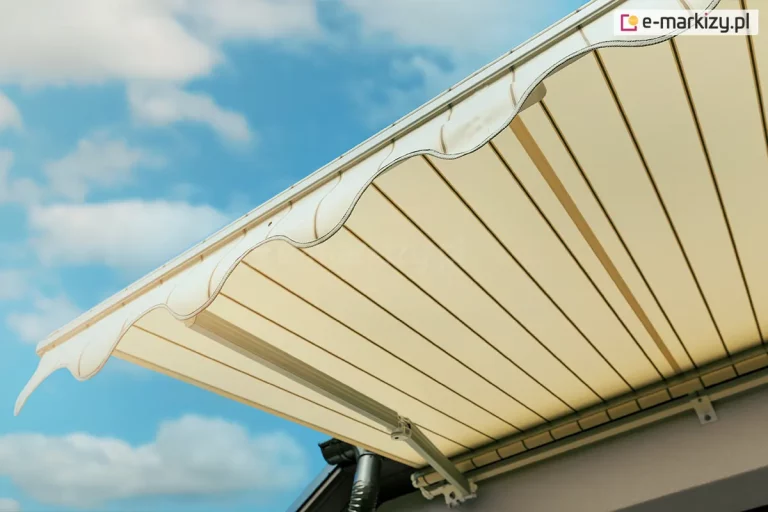 Zbliżenie na krawędź przednią i spód poszycia markizy Adagio, ukazujące detale wykończenia tkaniny oraz konstrukcję falbany. Materiał markizy ma kolor kremowy lub jasnobeżowy z subtelnymi, ciemniejszymi pionowymi pasami o różnej szerokości. Na krawędzi czołowej znajduje się dekoracyjna falbana o klasycznym, falistym kształcie, która została obszyta ciemniejszą lamówką dla wzmocnienia brzegu i podkreślenia formy. Tkanina jest precyzyjnie napięta na aluminiowym profilu przednim w kolorze białym. Pod poszyciem widoczny jest fragment białego ramienia przegubowego oraz rury nawojowej markizy Adagio, zamontowanej na jasnej elewacji budynku. W dolnej części kadru zarysowuje się ciemna rynna dachowa. Tło zdjęcia stanowi pogodne, jasnoniebieskie niebo z rozproszonymi, białymi chmurami, co podkreśla estetykę i lekkość wizualną osłony przeciwsłonecznej.