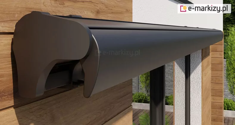 Elegancka, zamknięta kaseta markizy Palladio Selt w kolorze srebrzysty grafit, zamontowana na elewacji nowoczesnego budynku mieszkalnego. Obudowa ma opływowy, aerodynamiczny kształt i matowe wykończenie, a jej boczna pokrywa ściśle przylega do profilu frontowego, całkowicie chroniąc zwiniętą tkaninę przed czynnikami atmosferycznymi. System osłony jest przytwierdzony do ściany wykończonej poziomymi panelami z jasnego drewna o wyraźnym usłojeniu. W tle widoczny jest fragment białej, tynkowanej ściany z pionowymi przeszkleniami oraz zielone krzewy rosnące wzdłuż tarasu. Perspektywa zdjęcia z bliska podkreśla solidność wykonania konstrukcji oraz precyzyjne dopasowanie elementów mechanicznych, które po złożeniu tworzą estetyczną i minimalistyczną bryłę. Całość kompozycji oświetla naturalne światło dzienne, wydobywając fakturę drewna oraz gładkość metalowej obudowy.