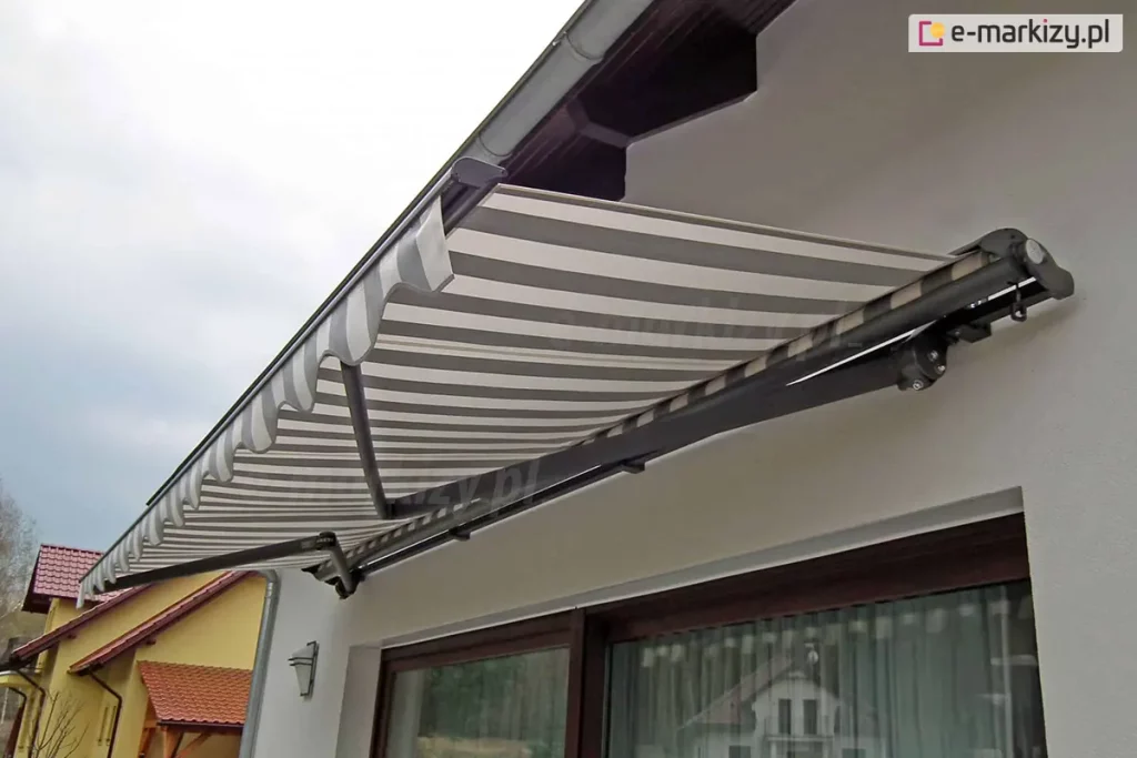 Zdjęcie przedstawia widok od dołu na w pełni rozwiniętą markizę Adagio, zamontowaną na białej elewacji nowoczesnego budynku. Poszycie markizy wykonane jest z wysokiej jakości tkaniny w poziome pasy w kolorach białym i jasnoszarym, która jest napięta na aluminiowej konstrukcji. Metalowy stelaż, obejmujący profil frontowy, ramiona przegubowe oraz rurę nawojową, został polakierowany proszkowo na ciemny, matowy kolor grafitowy. Po lewej stronie widoczna jest ozdobna falbana o falistym kształcie, wykończona białą lamówką. Markiza Adagio jest zamocowana tuż pod linią dachu, w pobliżu rynny, osłaniając duże przeszklenia tarasowe, w których odbija się sąsiednia zabudowa. Solidne ramiona przegubowe są widoczne w pełnym rozłożeniu, ukazując mechanizm łańcuchowy i przeguby łączące poszczególne sekcje wsporników. W tle, po lewej stronie kadru, widać fragmenty sąsiednich domów z żółtymi elewacjami i dachami pokrytymi czerwoną dachówką pod jasnym, zachmurzonym niebem.