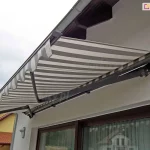 Zdjęcie przedstawia widok od dołu na w pełni rozwiniętą markizę Adagio, zamontowaną na białej elewacji nowoczesnego budynku. Poszycie markizy wykonane jest z wysokiej jakości tkaniny w poziome pasy w kolorach białym i jasnoszarym, która jest napięta na aluminiowej konstrukcji. Metalowy stelaż, obejmujący profil frontowy, ramiona przegubowe oraz rurę nawojową, został polakierowany proszkowo na ciemny, matowy kolor grafitowy. Po lewej stronie widoczna jest ozdobna falbana o falistym kształcie, wykończona białą lamówką. Markiza Adagio jest zamocowana tuż pod linią dachu, w pobliżu rynny, osłaniając duże przeszklenia tarasowe, w których odbija się sąsiednia zabudowa. Solidne ramiona przegubowe są widoczne w pełnym rozłożeniu, ukazując mechanizm łańcuchowy i przeguby łączące poszczególne sekcje wsporników. W tle, po lewej stronie kadru, widać fragmenty sąsiednich domów z żółtymi elewacjami i dachami pokrytymi czerwoną dachówką pod jasnym, zachmurzonym niebem.