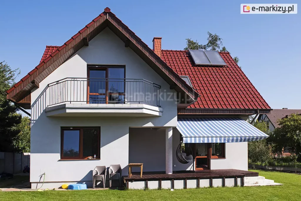 Zdjęcie prezentuje dwupiętrowy dom jednorodzinny z jasną elewacją i dwuspadowym dachem krytym czerwoną dachówką ceramiczną, na którym zamontowana jest markiza Mezzo mol. Dom posiada białe ściany, a stolarka okienna i drzwiowa wykonana jest w kolorze ciemnego brązu. Na wyższej kondygnacji znajduje się balkon z metalową, czarną balustradą o prostym wzorze. Na dachu widoczne są panele solarne oraz ceglany komin. Markiza Mezzo mol jest zamontowana na parterze, nad tarasem wykonanym z ciemnych desek kompozytowych lub drewnianych. Poszycie markizy ma wzór w szerokie i wąskie pasy w różnych odcieniach błękitu oraz bieli, co kontrastuje z ciepłą barwą dachu. Markiza jest w pełni rozłożona, rzucając wyraźny cień na część tarasu oraz znajdujący się pod nią czarny, wiszący fotel ogrodowy typu kokon. Taras jest lekko podwyższony względem otaczającego go, równo przystrzyżonego zielonego trawnika. Obok tarasu, przy ścianie budynku, stoją dwa szare krzesła ogrodowe z tworzywa sztucznego oraz mały stolik. W tle widoczne są zielone drzewa i fragmenty sąsiednich posesji, a całość sceny oświetlona jest ostrym, naturalnym światłem słonecznym w bezchmurny dzień.