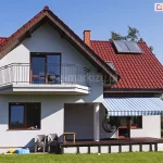 Zdjęcie prezentuje dwupiętrowy dom jednorodzinny z jasną elewacją i dwuspadowym dachem krytym czerwoną dachówką ceramiczną, na którym zamontowana jest markiza Mezzo mol. Dom posiada białe ściany, a stolarka okienna i drzwiowa wykonana jest w kolorze ciemnego brązu. Na wyższej kondygnacji znajduje się balkon z metalową, czarną balustradą o prostym wzorze. Na dachu widoczne są panele solarne oraz ceglany komin. Markiza Mezzo mol jest zamontowana na parterze, nad tarasem wykonanym z ciemnych desek kompozytowych lub drewnianych. Poszycie markizy ma wzór w szerokie i wąskie pasy w różnych odcieniach błękitu oraz bieli, co kontrastuje z ciepłą barwą dachu. Markiza jest w pełni rozłożona, rzucając wyraźny cień na część tarasu oraz znajdujący się pod nią czarny, wiszący fotel ogrodowy typu kokon. Taras jest lekko podwyższony względem otaczającego go, równo przystrzyżonego zielonego trawnika. Obok tarasu, przy ścianie budynku, stoją dwa szare krzesła ogrodowe z tworzywa sztucznego oraz mały stolik. W tle widoczne są zielone drzewa i fragmenty sąsiednich posesji, a całość sceny oświetlona jest ostrym, naturalnym światłem słonecznym w bezchmurny dzień.