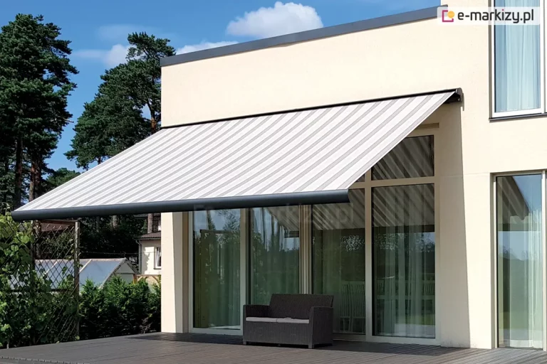 Zdjęcie prezentuje nowoczesną markizę tarasową Palladio Selt zainstalowaną na elewacji piętrowego budynku o minimalistycznej architekturze. Markiza posiada duży wysięg i jest pokryta tkaniną w szerokie, pionowe pasy w odcieniach bieli i jasnej szarości. Konstrukcja markizy, w tym kaseta i przednia belka, wykonana jest z metalu w kolorze antracytowym, co kontrastuje z jasną, kremową elewacją domu. Duży kąt nachylenia markizy zapewnia pełne zacienienie wyjścia na taras umieszczone w dużym przeszkleniu na fasadzie. Pod markizą znajduje się taras wykonany z ciemnobrązowych desek kompozytowych, na którym ustawiono pojedynczą, dwuosobową sofę z ciemnej plecionki z jasnym siedziskiem. Tło dla mebla stanowią duże, przeszklone drzwi tarasowe i okna, w których odbija się zieleń ogrodu. Po lewej stronie widoczne jest gęste ogrodzenie z siatki porośnięte roślinnością oraz wysokie sosny na tle błękitnego, lekko zachmurzonego nieba. Architektura budynku charakteryzuje się prostymi liniami i brakiem zbędnych zdobień, co podkreśla funkcjonalny i luksusowy charakter prezentowanej osłony przeciwsłonecznej.