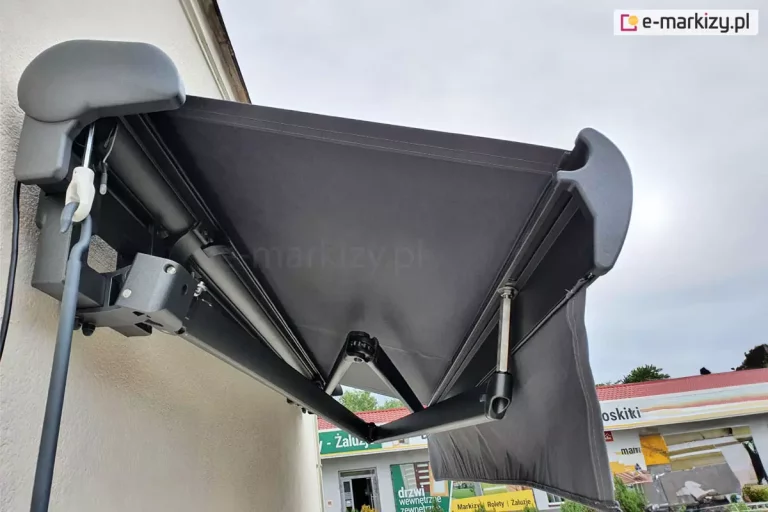 Zawieszona na ścianie budynku z tynkiem strukturalnym w kolorze białym markiza Palladio z grafitową konstrukcją oraz tkaniną i prostą falbaną w kolorze Antracita z widocznymi przy niewielkim rozsunięciu skrzyżowanymi ramionami, które składają się przy belce jedno nad drugim. Po lewej widoczny zaczep oczkowy z korbą do ręcznego sterowania.