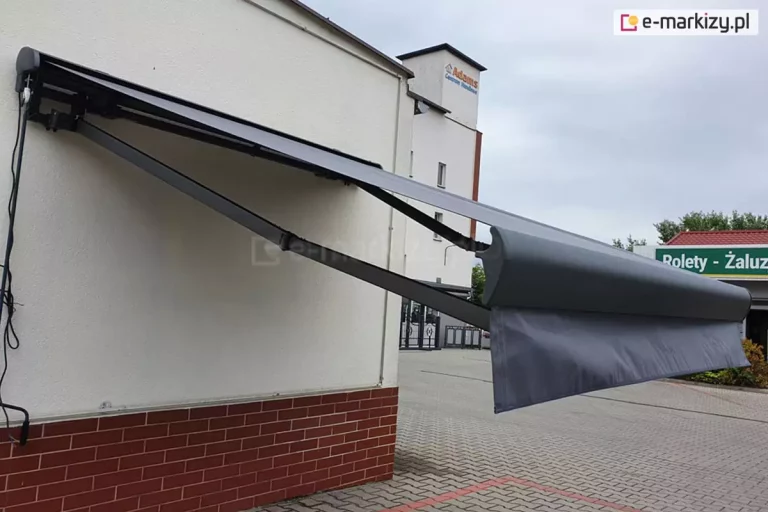 Grafitowa markiza Palladio Selt zamocowana na ścianie budynku z ceglaną podmurówką, markiza z ramionami krzyżowymi, z których lewy wysięgnik zainstalowany jest niżej niż prawy, dzięki czemu składają się one równolegle.