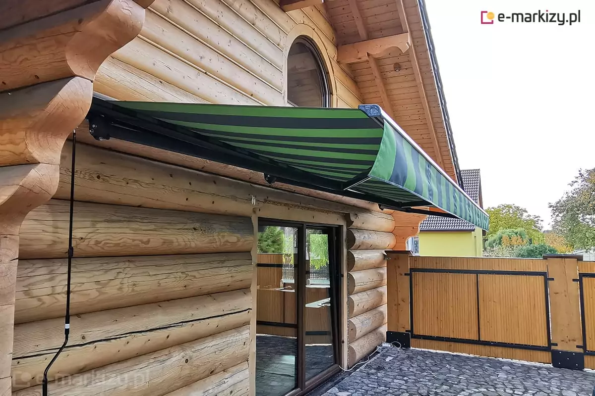 Zamocowana na ścianie chaty z bali markiza Adagio z czarną konstrukcją i poszyciem w zielono-czarne pasy osłania duże wyjście na taras wyłożony kostka brukową i ograniczony drewnianym płotem z pełnymi panelami chroniącymi przed spojrzeniami przechodniów.