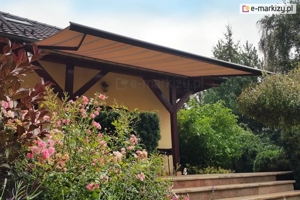 Przykład zastosowania markizy firmy Mol model Presto do osłony tarasu. Na zdjęciu widać markizę Presto zamontowaną na bocznej elewacji budynku jednorodzinnego pod okapem dachu. Poszycie markizy wykonane jest z tkaniny w odcieniu ciepłego beżu zdobionej regularnymi, poprzecznymi pasami w nieco ciemniejszym kolorze. Materiał jest napięty na solidnej, ciemnobrązowej konstrukcji aluminiowej, której belka frontowa oraz ramiona przegubowe są wyraźnie widoczne. Markiza osłania taras, którego konstrukcja opiera się na masywnych, drewnianych belkach o ciemnym wykończeniu, tworzących zadaszenie zintegrowane z elewacją domu w kolorze jasnego kremu. Na pierwszym planie, po lewej stronie, znajdują się obficie kwitnące krzewy róż o jasnoróżowych kwiatach, które częściowo przesłaniają widok na schody prowadzące na taras. Schody wykonane są z kamienia lub płytek w kolorze piaskowym. Po prawej stronie oraz w tle rozpościera się gęsty, wielopoziomowy ogród z różnorodnymi krzewami liściastymi i iglastymi, w tym widoczną brzozą o zwisających gałęziach. Na samym tarasie, przy balustradzie, można dostrzec małą figurkę białego kota. Dach budynku pokryty jest ciemną, grafitową dachówką, a niebo nad posesją jest jasne z delikatnymi rozmyciami chmur. Całość kompozycji ukazuje estetyczne i funkcjonalne dopasowanie osłony przeciwsłonecznej do architektury domu i otaczającej go roślinności.