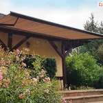 Przykład zastosowania markizy firmy Mol model Presto do osłony tarasu. Na zdjęciu widać markizę Presto zamontowaną na bocznej elewacji budynku jednorodzinnego pod okapem dachu. Poszycie markizy wykonane jest z tkaniny w odcieniu ciepłego beżu zdobionej regularnymi, poprzecznymi pasami w nieco ciemniejszym kolorze. Materiał jest napięty na solidnej, ciemnobrązowej konstrukcji aluminiowej, której belka frontowa oraz ramiona przegubowe są wyraźnie widoczne. Markiza osłania taras, którego konstrukcja opiera się na masywnych, drewnianych belkach o ciemnym wykończeniu, tworzących zadaszenie zintegrowane z elewacją domu w kolorze jasnego kremu. Na pierwszym planie, po lewej stronie, znajdują się obficie kwitnące krzewy róż o jasnoróżowych kwiatach, które częściowo przesłaniają widok na schody prowadzące na taras. Schody wykonane są z kamienia lub płytek w kolorze piaskowym. Po prawej stronie oraz w tle rozpościera się gęsty, wielopoziomowy ogród z różnorodnymi krzewami liściastymi i iglastymi, w tym widoczną brzozą o zwisających gałęziach. Na samym tarasie, przy balustradzie, można dostrzec małą figurkę białego kota. Dach budynku pokryty jest ciemną, grafitową dachówką, a niebo nad posesją jest jasne z delikatnymi rozmyciami chmur. Całość kompozycji ukazuje estetyczne i funkcjonalne dopasowanie osłony przeciwsłonecznej do architektury domu i otaczającej go roślinności.