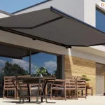 Nowoczesna markiza Dakar w kolorze ciemnoszarym zamontowana na elewacji białego, piętrowego budynku o minimalistycznej architekturze. Materiał poszycia markizy jest napięty i rozłożony nad przestronnym tarasem, chroniąc przed słońcem zestaw mebli ogrodowych. Konstrukcja wspornikowa markizy opiera się na bocznych ramionach przegubowych, które utrzymują tkaninę w pozycji poziomej z lekkim nachyleniem. Pod markizą znajduje się okrągły, ciemny stół oraz kilka drewnianych krzeseł z oparciami z poziomych listew. Przeszklone, duże drzwi tarasowe odbijają błękitne niebo z białymi chmurami oraz zieleń drzew znajdujących się w ogrodzie. Część ściany budynku pod markizą została wykończona panelami o teksturze jasnego drewna, co kontrastuje z białą fasadą i ciemnym kolorem osłony. Z prawej strony tarasu widoczny jest fragment drewnianego wózka barowego oraz drugie, duże przeszklenie na piętrze budynku. Cała scena przedstawia funkcjonalne wykorzystanie markizy Dakar w aranżacji nowoczesnej strefy wypoczynkowej na świeżym powietrzu.