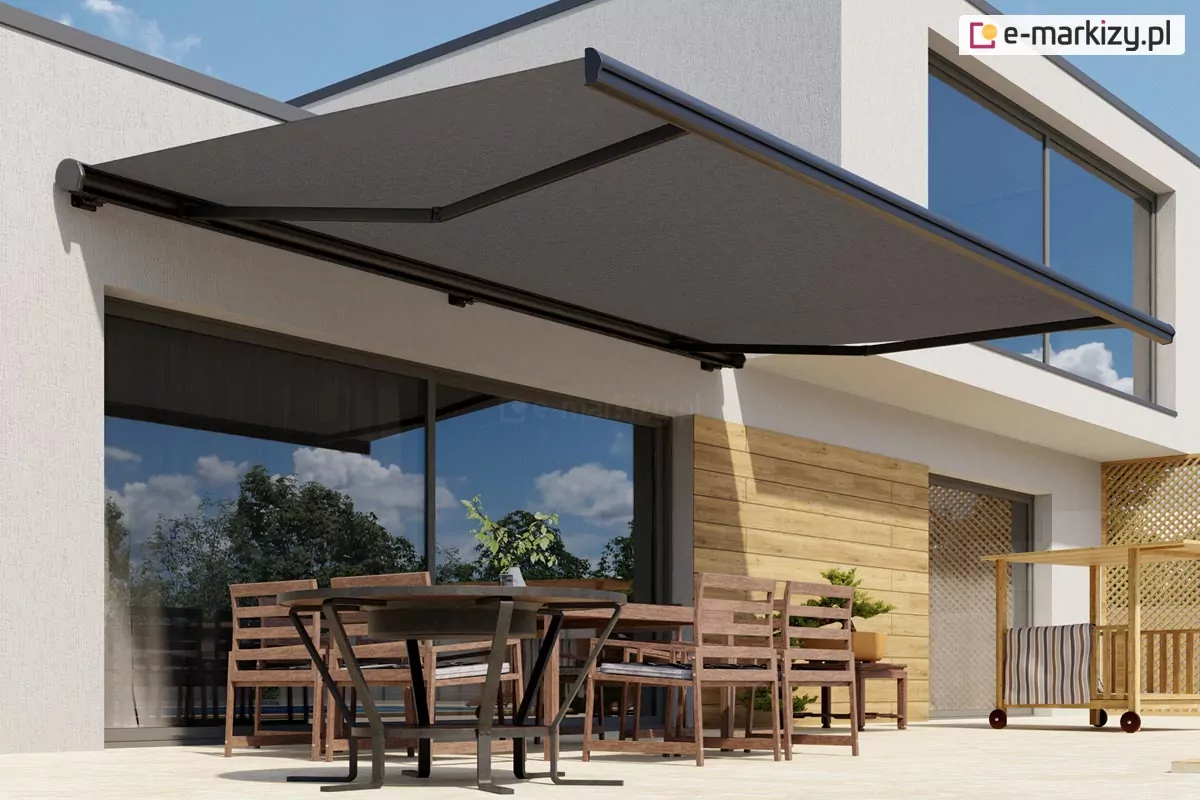 Nowoczesna markiza Dakar w kolorze ciemnoszarym zamontowana na elewacji białego, piętrowego budynku o minimalistycznej architekturze. Materiał poszycia markizy jest napięty i rozłożony nad przestronnym tarasem, chroniąc przed słońcem zestaw mebli ogrodowych. Konstrukcja wspornikowa markizy opiera się na bocznych ramionach przegubowych, które utrzymują tkaninę w pozycji poziomej z lekkim nachyleniem. Pod markizą znajduje się okrągły, ciemny stół oraz kilka drewnianych krzeseł z oparciami z poziomych listew. Przeszklone, duże drzwi tarasowe odbijają błękitne niebo z białymi chmurami oraz zieleń drzew znajdujących się w ogrodzie. Część ściany budynku pod markizą została wykończona panelami o teksturze jasnego drewna, co kontrastuje z białą fasadą i ciemnym kolorem osłony. Z prawej strony tarasu widoczny jest fragment drewnianego wózka barowego oraz drugie, duże przeszklenie na piętrze budynku. Cała scena przedstawia funkcjonalne wykorzystanie markizy Dakar w aranżacji nowoczesnej strefy wypoczynkowej na świeżym powietrzu.
