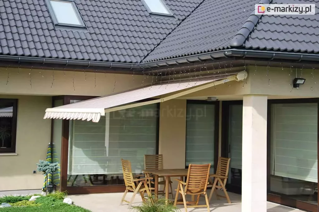 Markiza w kasecie Dakar Aluprof zamocowana do ściany pod podbitką dachu pokrytego grafitową dachówką. Wysunięte poszycie w białe i jasnoczerwone pasy zacienia przestrzeń niewielkiego tarasu, na którym ustawiono drewniany stół z czterema krzesłami z drewna o wysokich oparciach oraz duże okna tarasowe przesłonięte dodatkowo opuszczonymi roletami rzymskimi o białej tkaninie. Do podbitki dachu przymocowane są światełka choinkowe. Obok tarasu widoczne małe drzewko rosnące na skrawku trawnika, a za nim zwinięty parasol ogrodowy w zielone i niebieskie pasy.