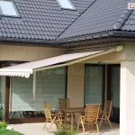 Markiza w kasecie Dakar Aluprof zamocowana do ściany pod podbitką dachu pokrytego grafitową dachówką. Wysunięte poszycie w białe i jasnoczerwone pasy zacienia przestrzeń niewielkiego tarasu, na którym ustawiono drewniany stół z czterema krzesłami z drewna o wysokich oparciach oraz duże okna tarasowe przesłonięte dodatkowo opuszczonymi roletami rzymskimi o białej tkaninie. Do podbitki dachu przymocowane są światełka choinkowe. Obok tarasu widoczne małe drzewko rosnące na skrawku trawnika, a za nim zwinięty parasol ogrodowy w zielone i niebieskie pasy.