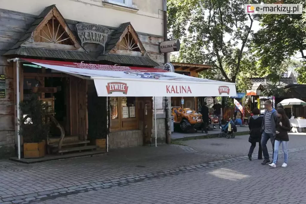 Markiza reklamowa z nadrukami na poszyciu i falbanie stanowi zacienienie wejścia do restauracji Kalina. Markiza Australia umieszczona nad wejściem do restauracji zacienia ogródek z kostki brukowej. Na deptaku widoczni także spacerujący turyści.