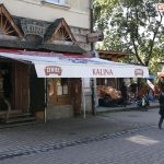 Markiza reklamowa z nadrukami na poszyciu i falbanie stanowi zacienienie wejścia do restauracji Kalina. Markiza umieszczona nad wejściem do restauracji zacienia ogródek z kostki brukowej. Na deptaku widoczni także spacerujący turyści.