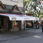 Markiza reklamowa z nadrukami na poszyciu i falbanie stanowi zacienienie wejścia do restauracji Kalina. Markiza Australia umieszczona nad wejściem do restauracji zacienia ogródek z kostki brukowej. Na deptaku widoczni także spacerujący turyści.