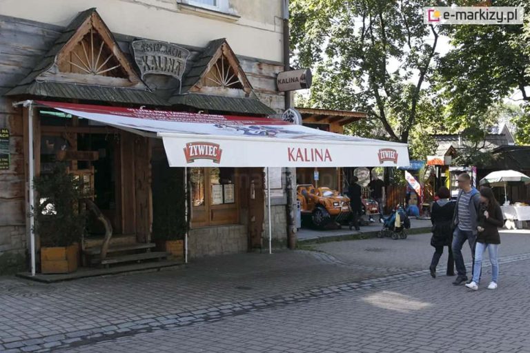 Markiza reklamowa z nadrukami na poszyciu i falbanie stanowi zacienienie wejścia do restauracji Kalina. Markiza umieszczona nad wejściem do restauracji zacienia ogródek z kostki brukowej. Na deptaku widoczni także spacerujący turyści.