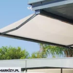 Zdjęcie prezentuje nowoczesny system osłonowy markiza Moderno zamontowany pod stropem balkonu lub tarasu, co obrazuje wariant montażu sufitowego. Markiza jest w pełni rozwinięta, a jej konstrukcja, obejmująca ramiona przegubowe i profil frontowy, wykonana jest z aluminium w eleganckim, ciemnym kolorze antracytowym. Poszycie stanowi jasna, beżowa tkanina o gładkiej strukturze, która jest mocno napięta, tworząc skuteczną barierę przed słońcem. W dolnej części kadru widoczna jest szklana balustrada nowoczesnego tarasu, a tło wypełnia gęsta, soczyście zielona roślinność ogrodowa z drzewami iglastymi i liściastymi pod bezchmurnym, błękitnym niebem. Minimalistyczny profil markizy bez falbany podkreśla współczesny design produktu, idealnie pasujący do luksusowych apartamentów i rezydencji.