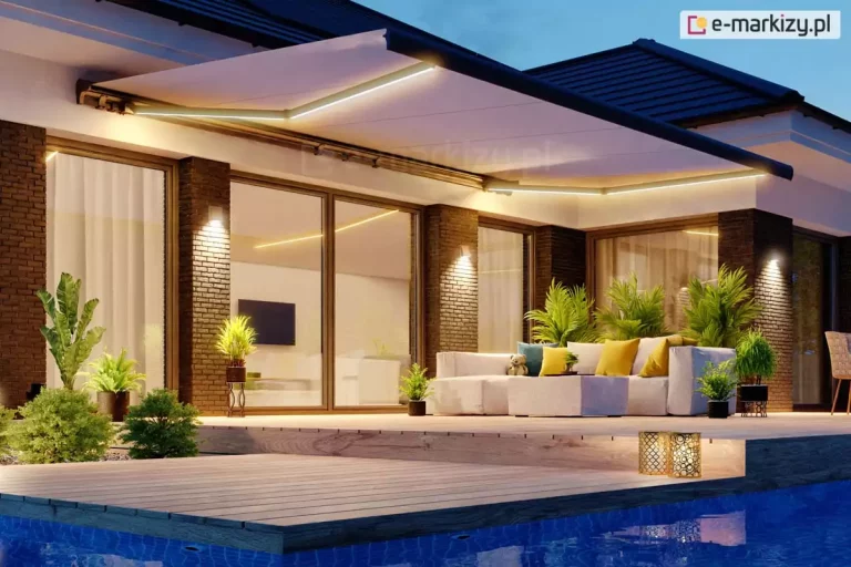 Zdjęcie prezentuje luksusową aranżację tarasu przy nowoczesnym domu jednorodzinnym w porze wieczornej. Centralnym elementem jest duża, rozłożona markiza tarasowa Palladio z poszyciem w kolorze białym lub bardzo jasnoszarym, wyposażona w zintegrowane oświetlenie LED wzdłuż ramion przegubowych. Ciepłe światło z markizy oraz kinkietów na elewacji oświetla strefę wypoczynkową, na którą składa się duży, narożny modułowy narożnik w jasnym kolorze z dekoracyjnymi żółtymi i niebieskimi poduszkami. Taras wykonany jest z drewnianych desek o wyraźnym usłojeniu i graniczy bezpośrednio z podświetlonym basenem o intensywnie niebieskiej wodzie. Elewacja budynku charakteryzuje się połączeniem ciemnej cegły klinkierowej z dużymi, panoramicznymi przeszkleniami, przez które widać nowocześnie urządzone wnętrze salonu. Na tarasie rozmieszczono liczne rośliny doniczkowe, w tym palmy i ozdobne krzewy, które nadają przestrzeni egzotyczny charakter. W prawym dolnym rogu, przy krawędzi basenu, znajdują się dwie dekoracyjne, ażurowe latarnie emitujące złote światło. Całość kompozycji utrzymana jest w eleganckiej, relaksującej atmosferze, podkreślonej przez kontrast między ciepłym oświetleniem a ciemniejącym, wieczornym niebem.