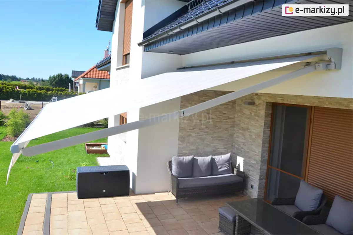 Szerokie ujęcie tarasu nowoczesnego domu jednorodzinnego, na którym zamontowana została rozłożona markiza Jamaica. Konstrukcja osłony przeciwsłonecznej opiera się na dwóch solidnych ramionach przegubowych wykonanych z aluminium lakierowanego na kolor biały, które podtrzymują napięty materiał poszycia. Tkanina w kolorze jasnokremowym lub białym z delikatnymi, poziomymi pasami skutecznie zacienia przestrzeń wypoczynkową, chroniąc meble ogrodowe przed bezpośrednim promieniowaniem słonecznym. Markiza Jamaica jest zamontowana do białej elewacji budynku, tuż pod linią okapu dachu pokrytego ciemną dachówką wyposażona została w dodatkową opcjonalną półkasetę - daszek markizowy. Pod markizą znajduje się zaaranżowana strefa relaksu z meblami z technorattanu w kolorze ciemnoszarym, w tym sofa z miękkimi, popielatymi poduszkami oraz fotele przy ciemnym stole. Ściana tarasu została wykończona dekoracyjnym kamieniem o beżowo-szarej kolorystyce, co harmonizuje z jasną nawierzchnią wykonaną z kostki brukowej. W tle widoczny jest zadbany, zielony trawnik, czarna skrzynia ogrodowa oraz fragment sąsiedniej zabudowy z dachami o czerwonej dachówce. Całość kompozycji pokazuje praktyczne zastosowanie markizy Jamaica jako estetycznego i funkcjonalnego elementu wyposażenia nowoczesnego tarasu, który zwiększa komfort użytkowania przestrzeni zewnętrznej w słoneczne dni.