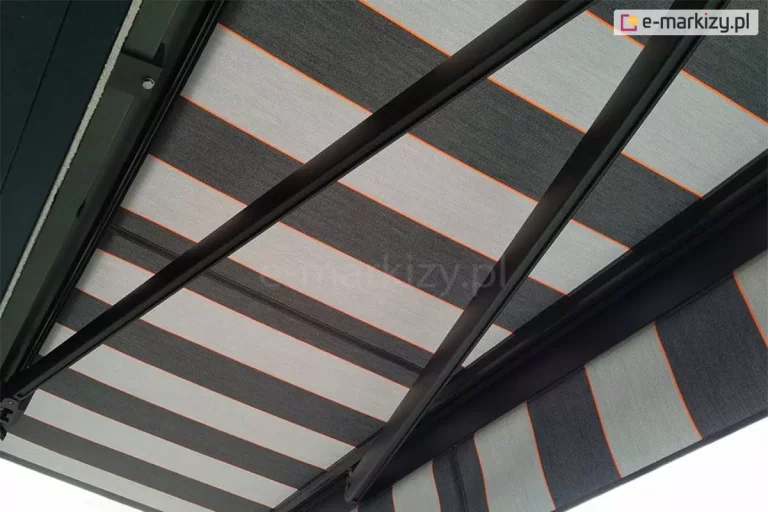 Widok markizy Palladio od spodu od strony częściowo wysuniętych ramion w kolorze czarnym z rozwiniętym nad nimi poszyciem w jasne i ciemne szare pasy przeplatane wąskimi czerwonymi liniami. Po prawej stronie widoczna prosta falbana wykonana z tej samej tkaniny, co poszycie.