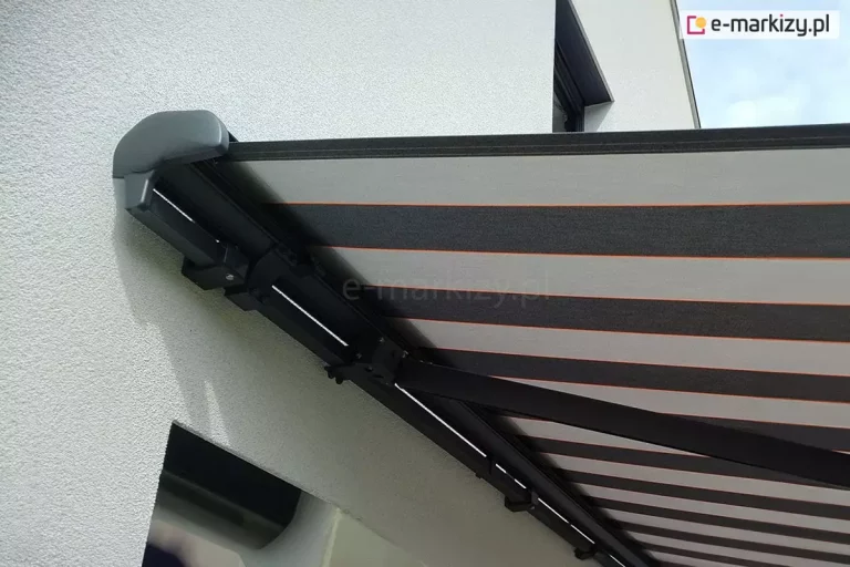 Markiza tarasowa Palladio z konstrukcją w kolorze srebrzysty grafit zamocowana na ścianie z białym tynkiem. Rozwinięte poszycie markizy w pasy o dwóch odcieniach szarości i wąskimi liniami w kolorze czerwonym pomiędzy nimi zacienia przestrzeń pod markizą.