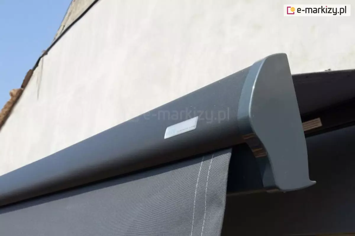 Belka przednia markizy Dakar z widoczną po prawej stronie zaślepką z pcv w kolorze konstrukcji srebrzysty grafit. Z belki wysunięta falbana w kolorze grafitowym z jasnoszarym przeszyciem. Ponad belką widoczne zwieńczenie dachu i niebo.