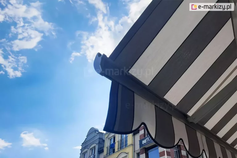 Widok belki przedniej markizy Palladio w kolorze brązowym z poszyciem i falowaną falbaną w pasy kremowe i brązowe na tle kamienic i pięknego, pogodnego błękitnego nieba z kilkoma białymi obłokami.