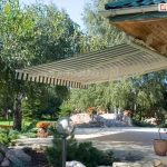 Zdjęcie prezentuje szerokie ujęcie tarasu przydomowego z rozłożoną markizą tarasową, która zapewnia cień nad strefą wypoczynkową. Markiza Australia posiada poszycie w klasyczne, pionowe pasy w kolorze białym i ciemnozielonym, zakończone ozdobną falbaną o tym samym wzorze. Konstrukcja wsporcza markizy została zamontowana bezpośrednio do krokwi dachowych pod okapem budynku, co pozwala na optymalne wykorzystanie przestrzeni bez konieczności montażu ściennego. Dom charakteryzuje się elewacją wykonaną z jasnego, nieregularnego kamienia dekoracyjnego oraz dachem pokrytym ciemnozieloną blachodachówką, wyposażonym w białe rynny PCV. Pod markizą znajduje się wybrukowany jasną kostką taras, na którym ustawiono donice z pomarańczowymi kwiatami oraz dekoracyjną lampę ogrodową w kształcie kuli. W tle widoczny jest starannie zagospodarowany ogród z gęstą roślinnością, wysokimi drzewami liściastymi, brzozą oraz skalniakiem wykonanym z dużych głazów. Całość kompozycji przedstawia praktyczne zastosowanie osłony przeciwsłonecznej w przydomowej architekturze krajobrazu w słoneczny dzień.