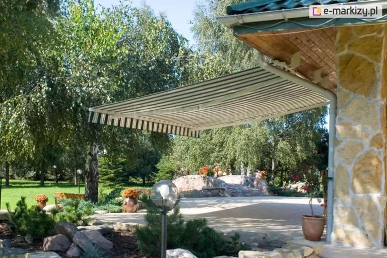 Zdjęcie prezentuje szerokie ujęcie tarasu przydomowego z rozłożoną markizą tarasową, która zapewnia cień nad strefą wypoczynkową. Markiza Australia posiada poszycie w klasyczne, pionowe pasy w kolorze białym i ciemnozielonym, zakończone ozdobną falbaną o tym samym wzorze. Konstrukcja wsporcza markizy została zamontowana bezpośrednio do krokwi dachowych pod okapem budynku, co pozwala na optymalne wykorzystanie przestrzeni bez konieczności montażu ściennego. Dom charakteryzuje się elewacją wykonaną z jasnego, nieregularnego kamienia dekoracyjnego oraz dachem pokrytym ciemnozieloną blachodachówką, wyposażonym w białe rynny PCV. Pod markizą znajduje się wybrukowany jasną kostką taras, na którym ustawiono donice z pomarańczowymi kwiatami oraz dekoracyjną lampę ogrodową w kształcie kuli. W tle widoczny jest starannie zagospodarowany ogród z gęstą roślinnością, wysokimi drzewami liściastymi, brzozą oraz skalniakiem wykonanym z dużych głazów. Całość kompozycji przedstawia praktyczne zastosowanie osłony przeciwsłonecznej w przydomowej architekturze krajobrazu w słoneczny dzień.